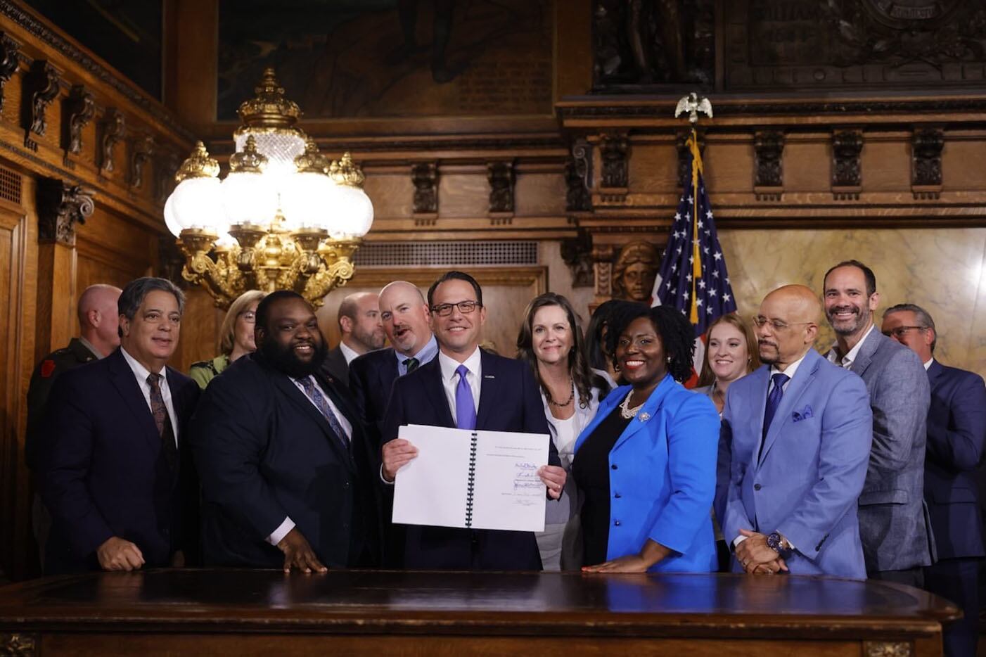 A group of people in business suits stands together for a portrait in a State Capitol room.