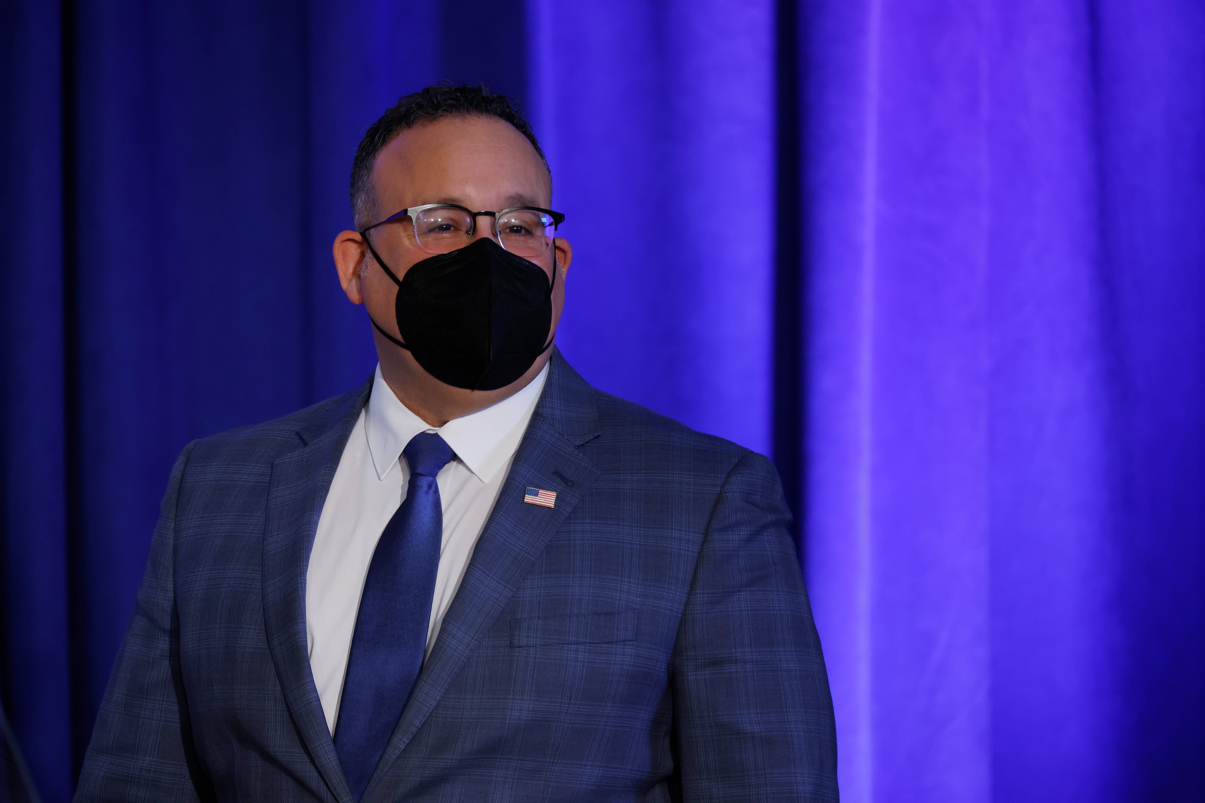 Secretary Miguel Cardona stands against a blue background, wearing glasses, a black mask, and a blue checkered suit.