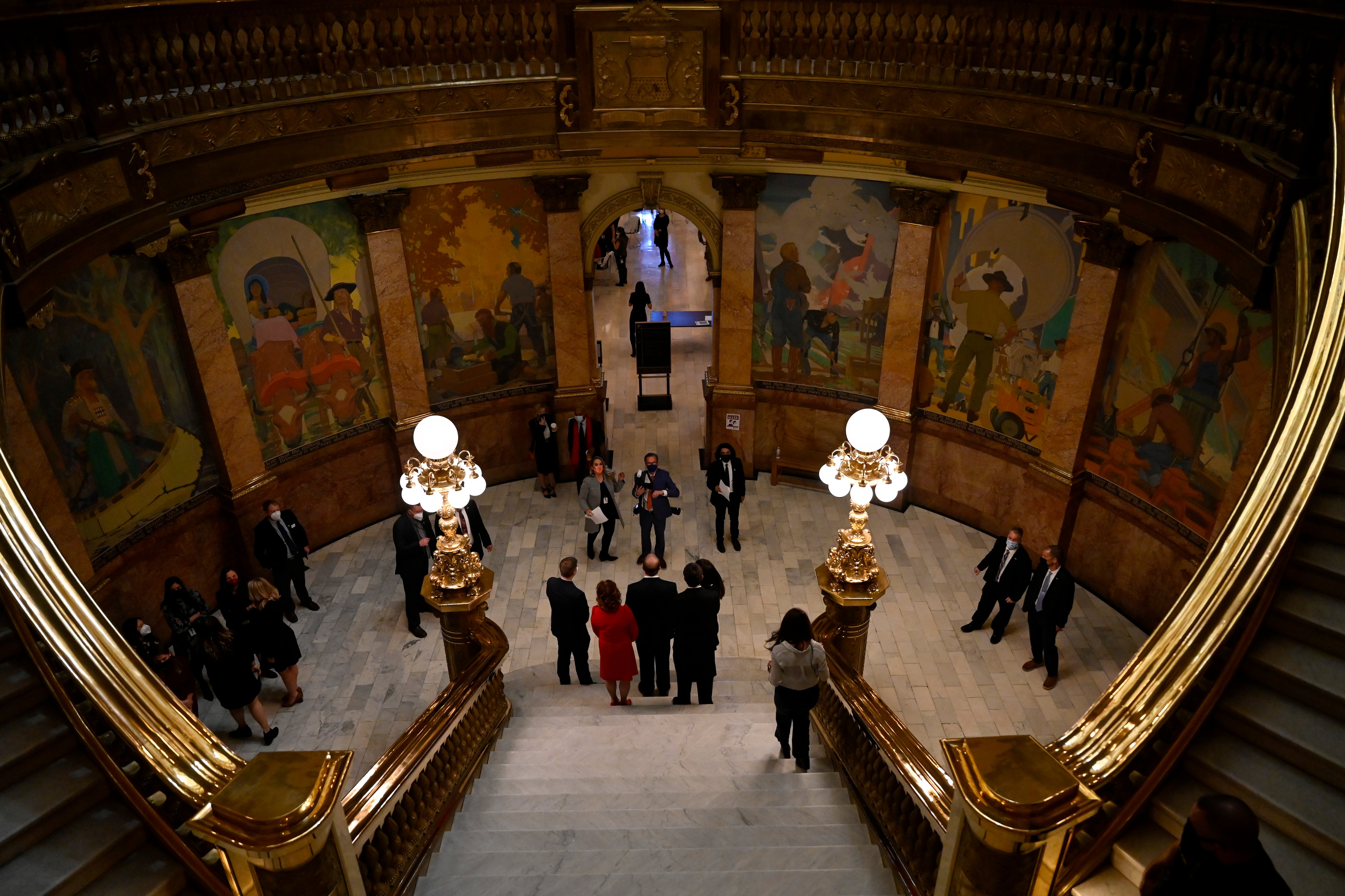 Several men and women stand at the bottom of grey steps and gold bannisters in the dimly lit Colorado Capitol.