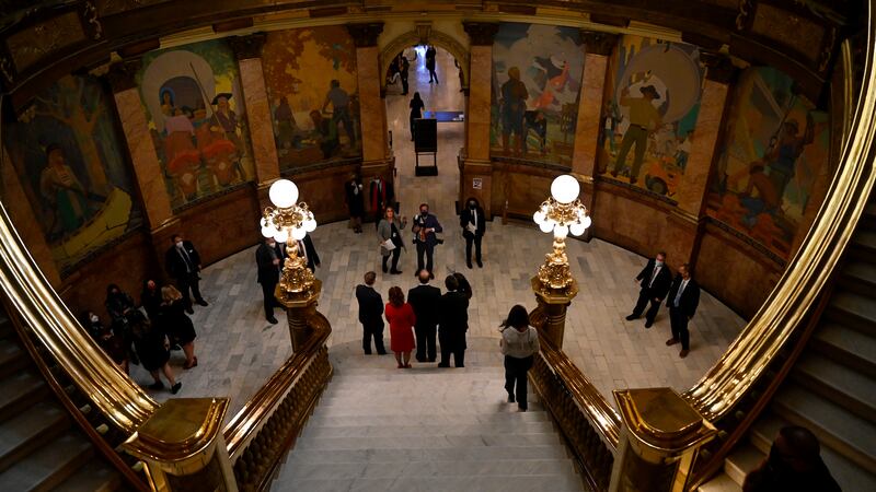 Several men and women stand at the bottom of grey steps and gold bannisters in the dimly lit Colorado Capitol.