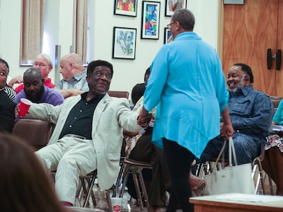 Man wearing a beige suit and black shirt shakes a woman’s hand while sitting