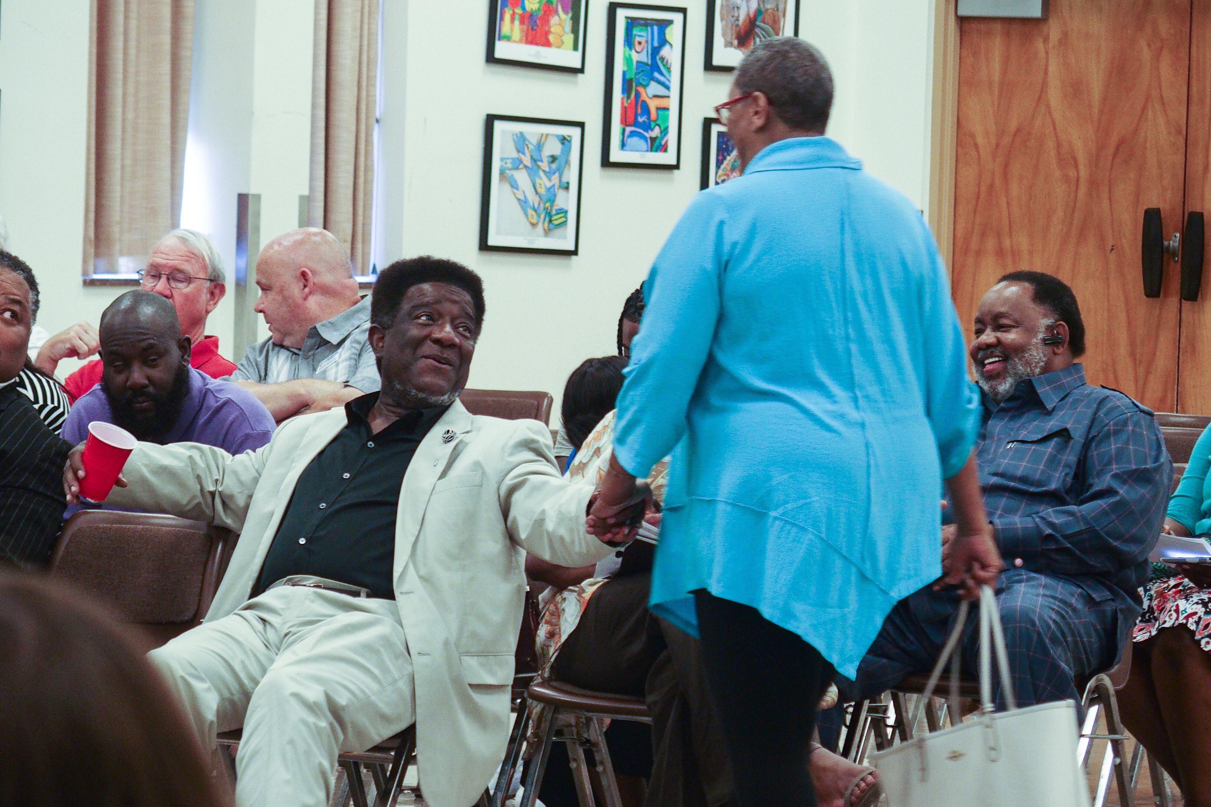 Man wearing a beige suit and black shirt shakes a woman’s hand while sitting
