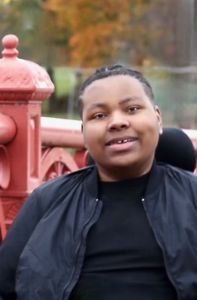 A headshot of a teenage boy with braided hair. He wears a black jacket and a black t-shirt.