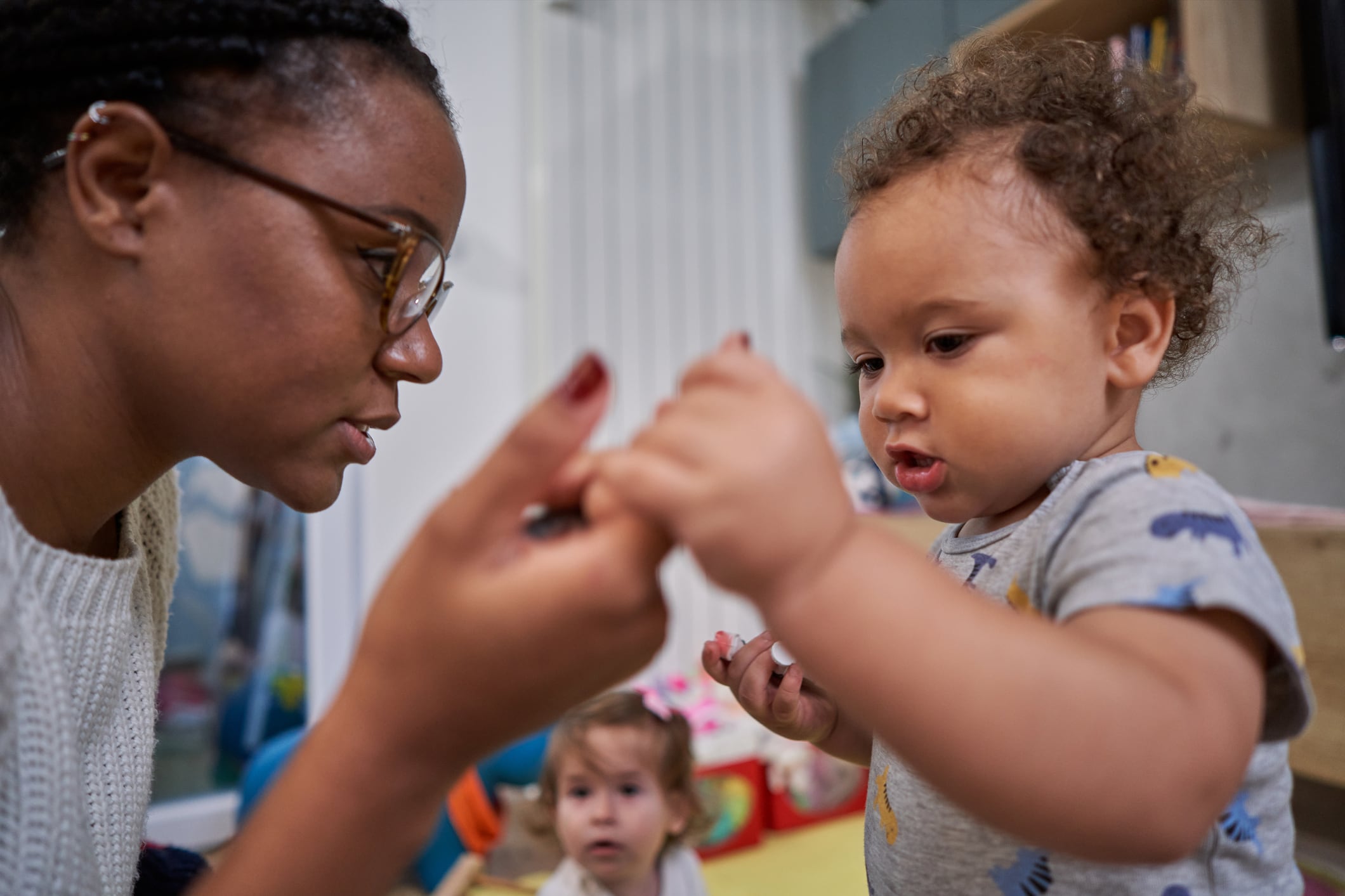 A young child holds the hand of a caregiver as another young child looks on in the background.