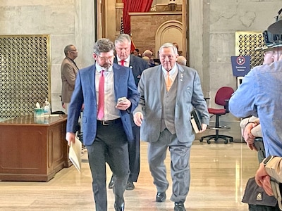 Two men wearing suits walk side by side in a large room with wood floors.