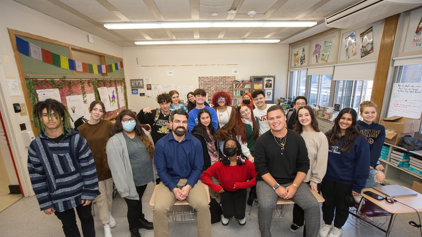 Twenty students assembled in a group in a classroom, with two adult male teachers sitting on desks directly in front of them.