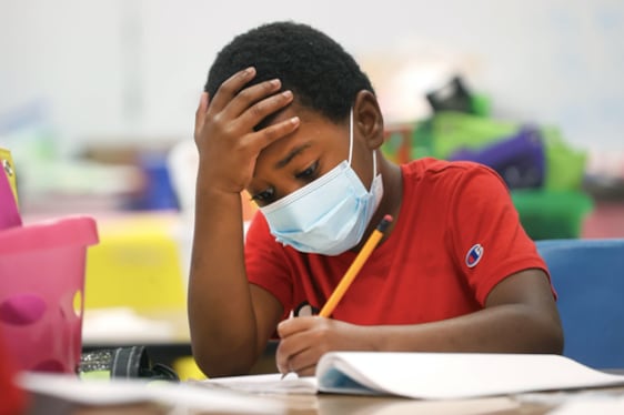 A young boy wearing a red shirt and a mask writes in a packet at his desk.