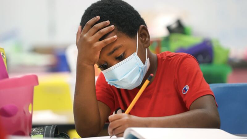 A young boy wearing a red shirt and a mask writes in a packet at his desk.
