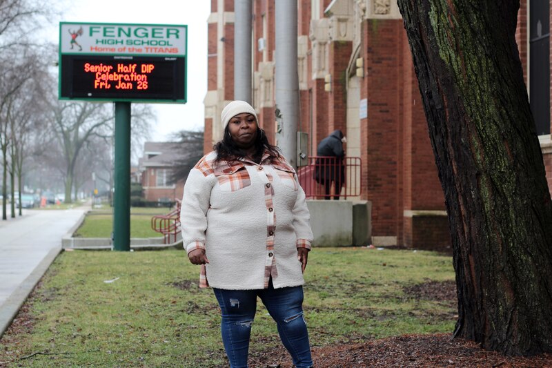 A woman with long dark hair and wearing a white jacket and tan hat stands near a tree and school building with the school sign in the background.