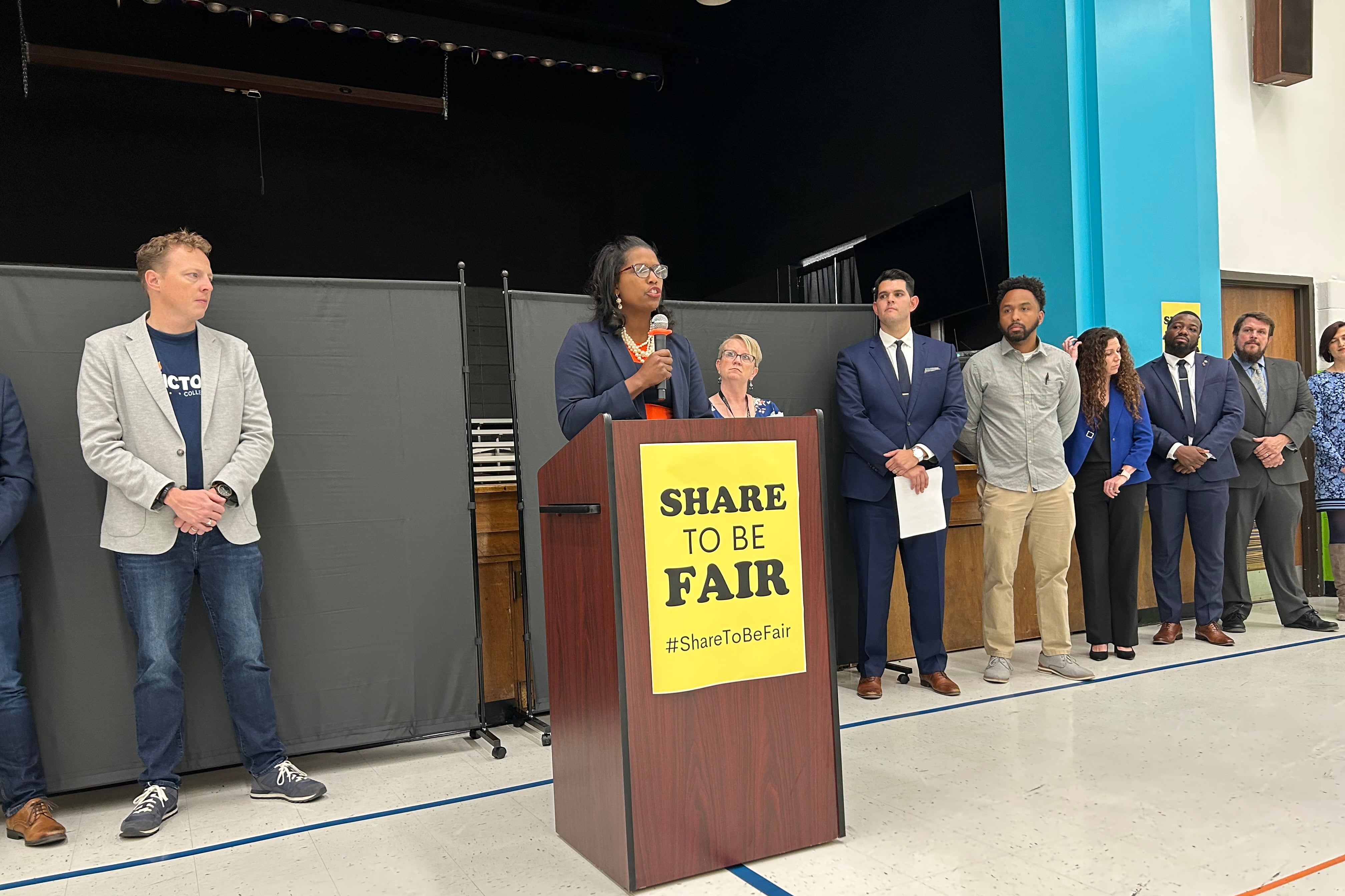 A woman holding a microphone stands behind a podium with a yellow sign on it that reads, “share to be fair.”
