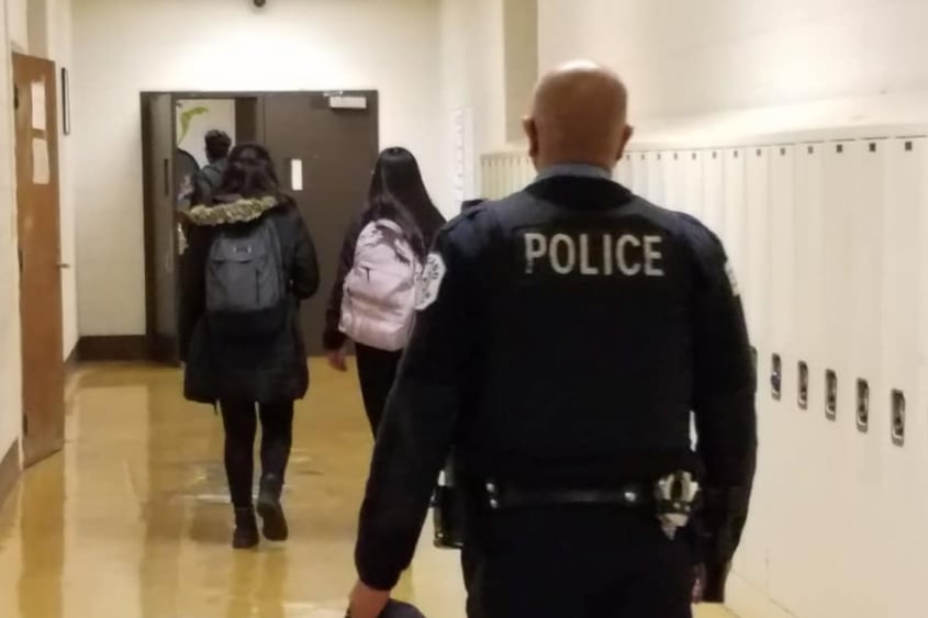 A policeman with POLICE written on the back of his uniform walks down a hallway lined with lockers and with two students walking in front of him.