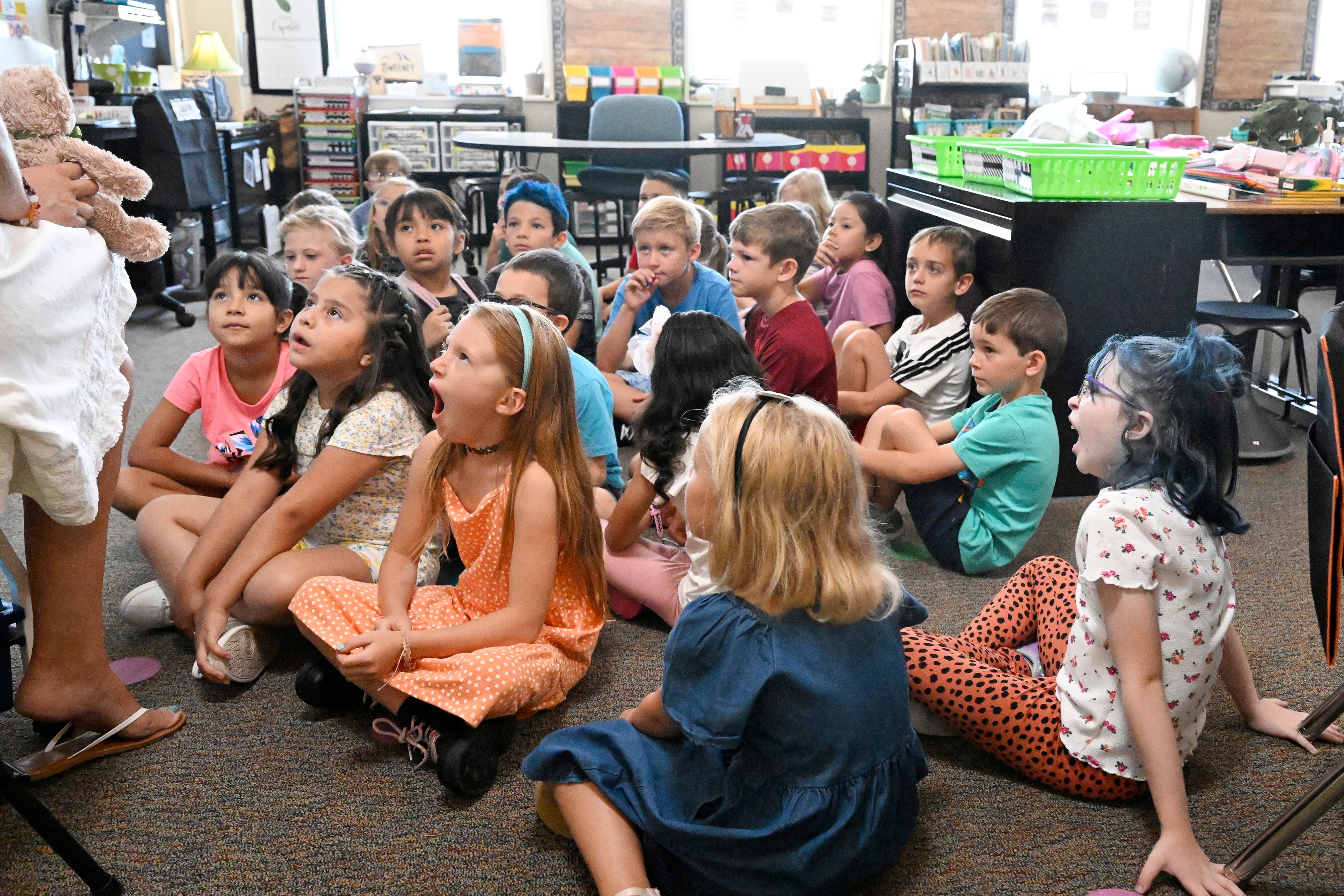 A classroom full of young students sit on the ground facing a teacher who is partially visible and desks in the background.
