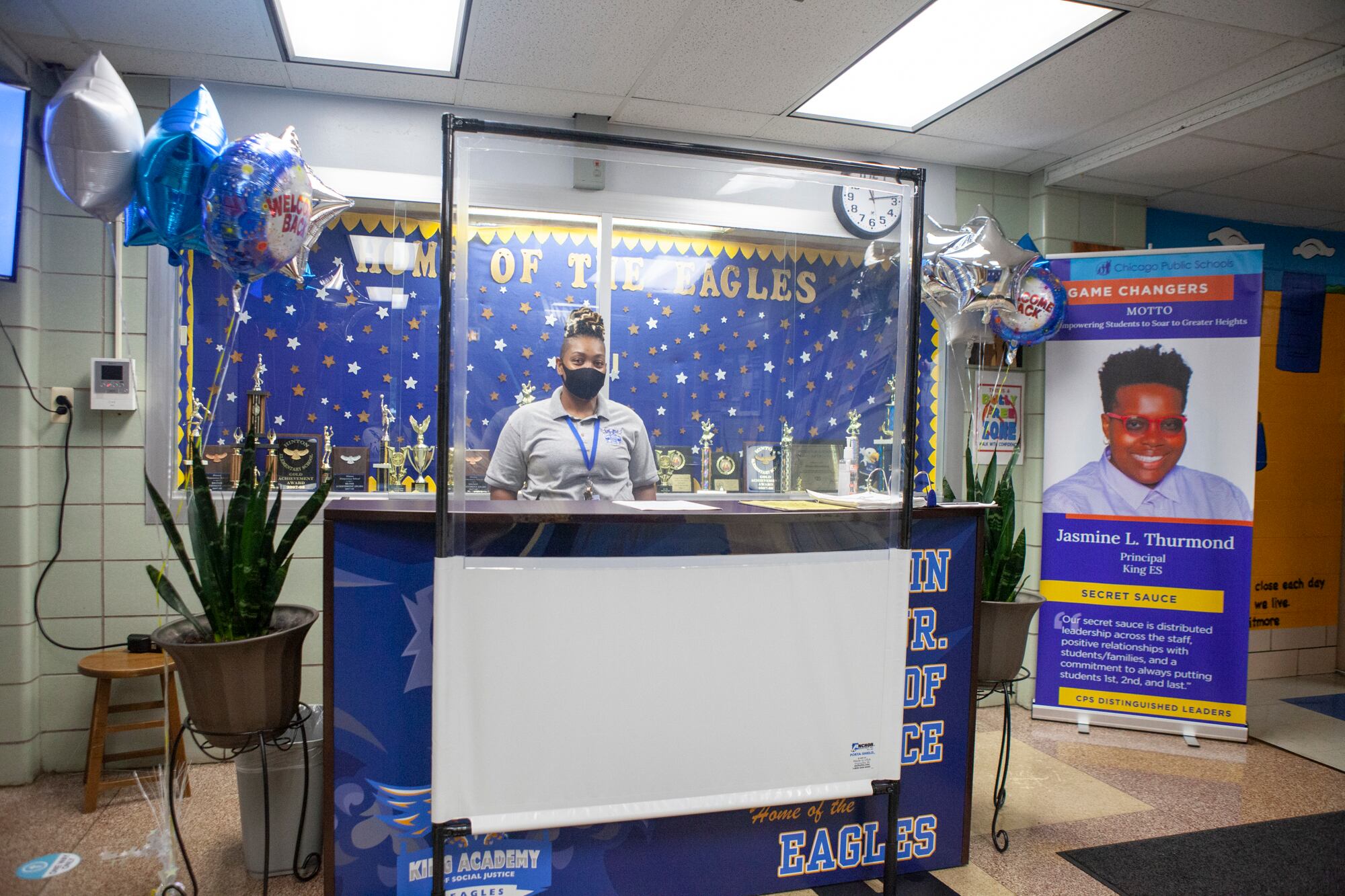 A health screening station with balloons that read welcome back and a man wearing a mask and behind a clear barrier.