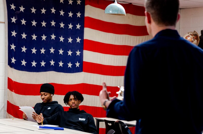 Two high school students sit at a desk while a teacher talks with them with a giant American flag in the background.