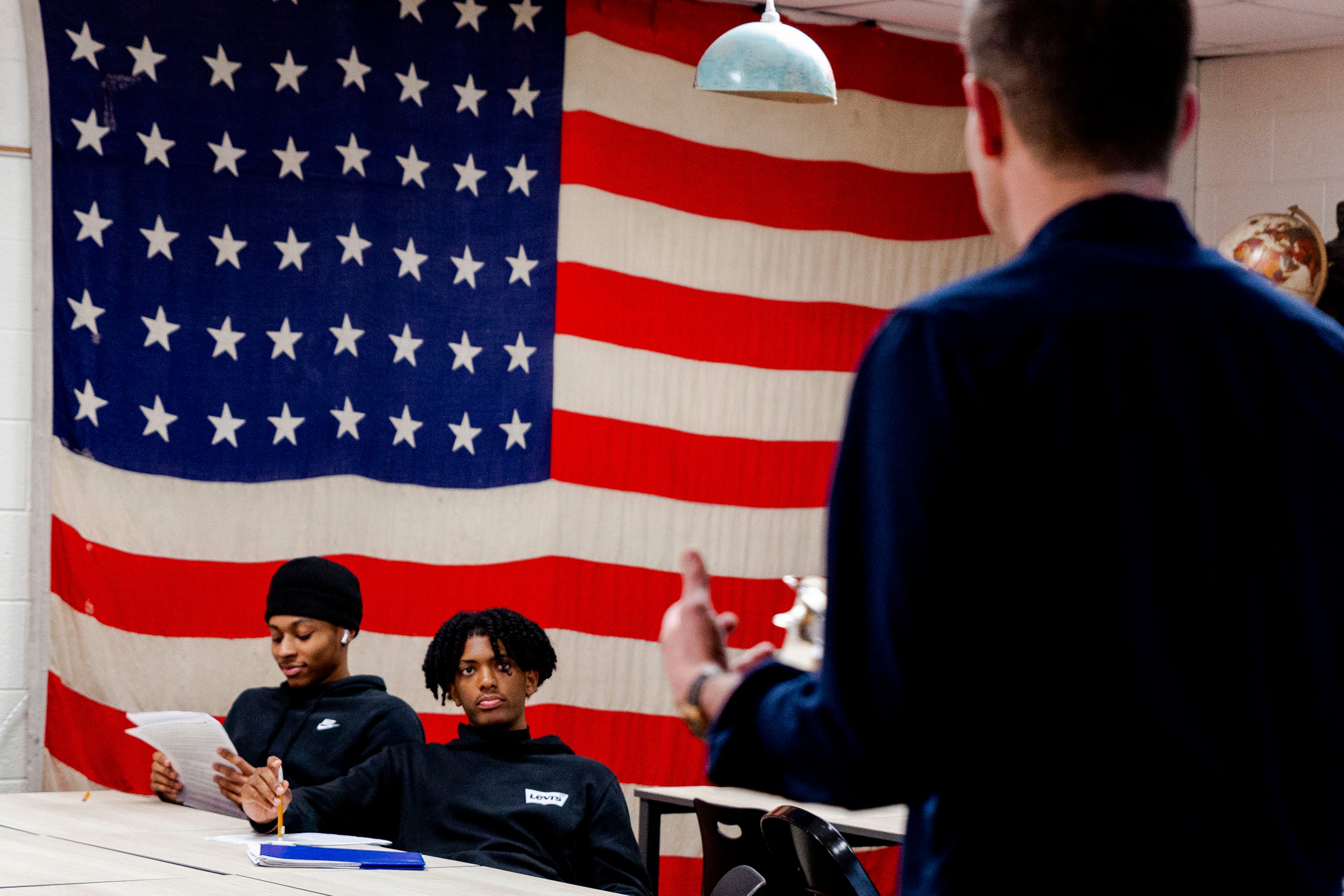 Two high school students sit at a desk while a teacher talks with them with a giant American flag in the background.