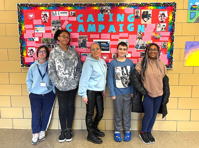 Four students and a principal stand in front of a bulletin board.