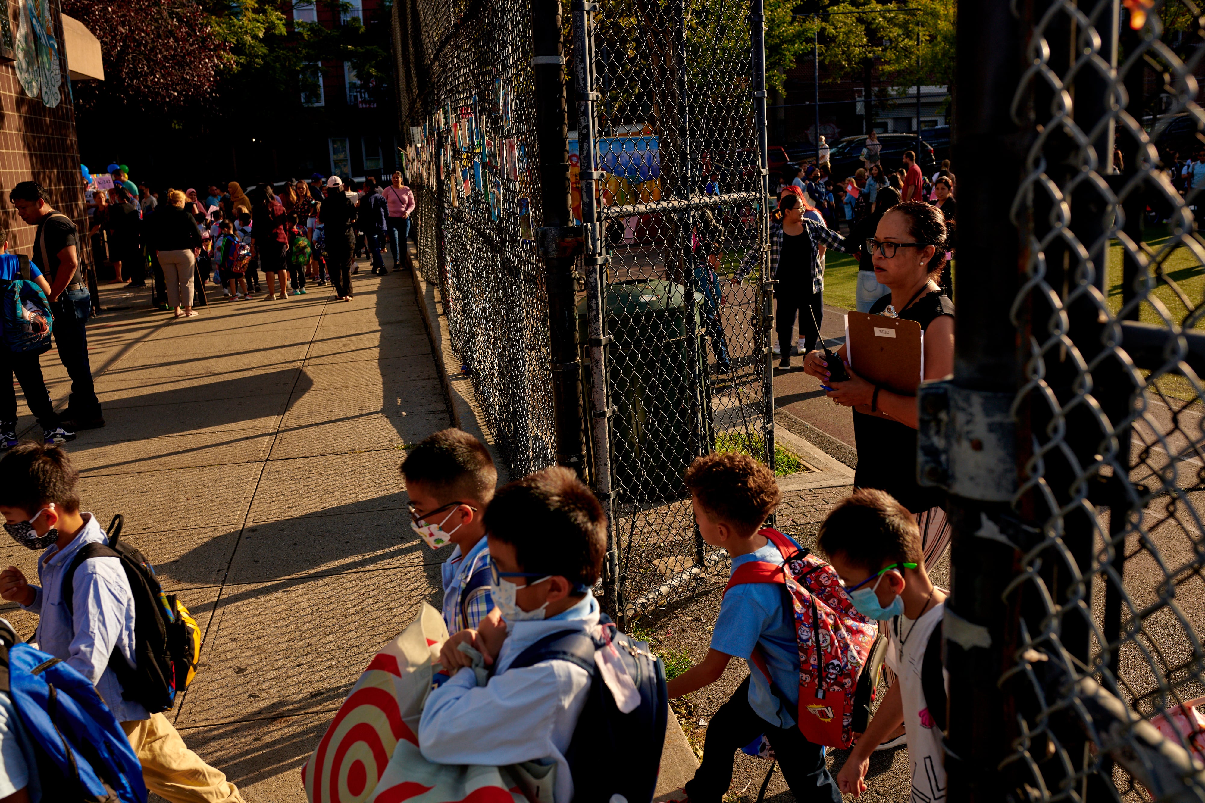 A woman in a black dress stands at a chain link fence around a school buildings as children, some masked, stream onto the sidewalk.
