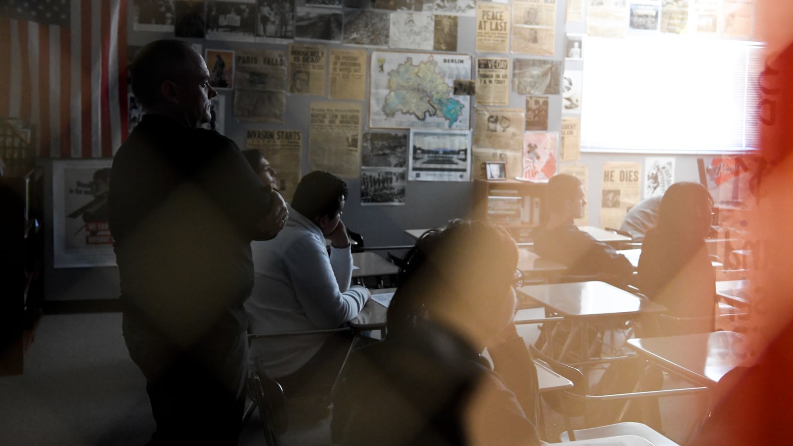 Several students sit at desks in a classroom while a teacher stands, with hazy lighting.