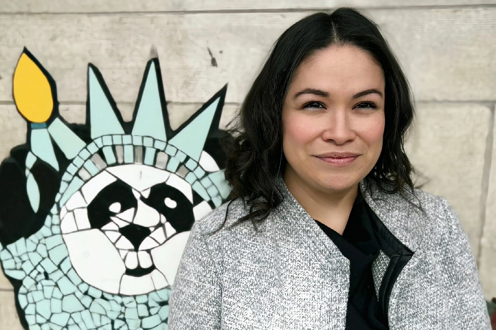 A smiling woman in a grey coat stands before a mosaic featuring the Statue of Liberty.