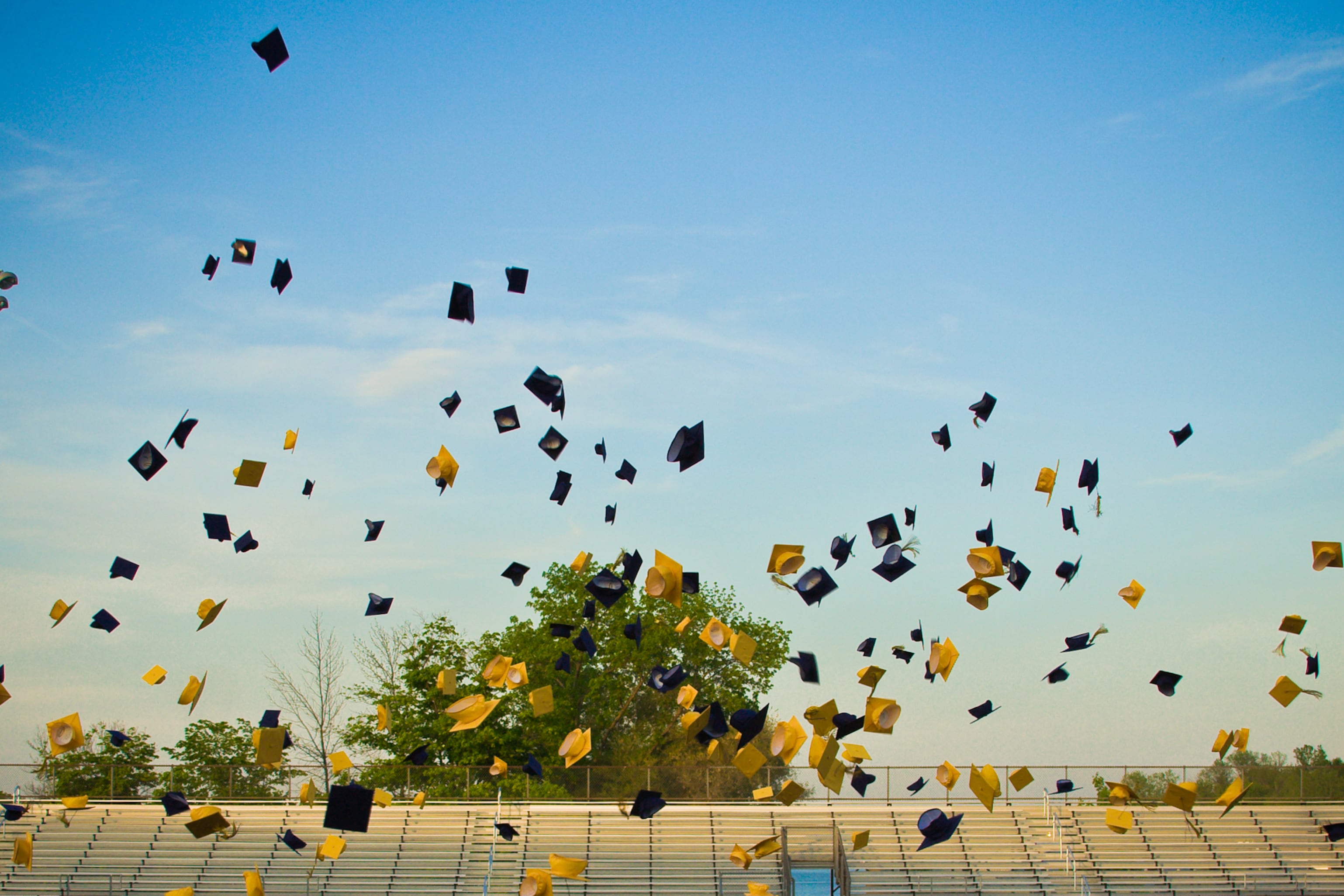 Blue and yellow graduation caps are in the air with a stadium and and lights in the background.