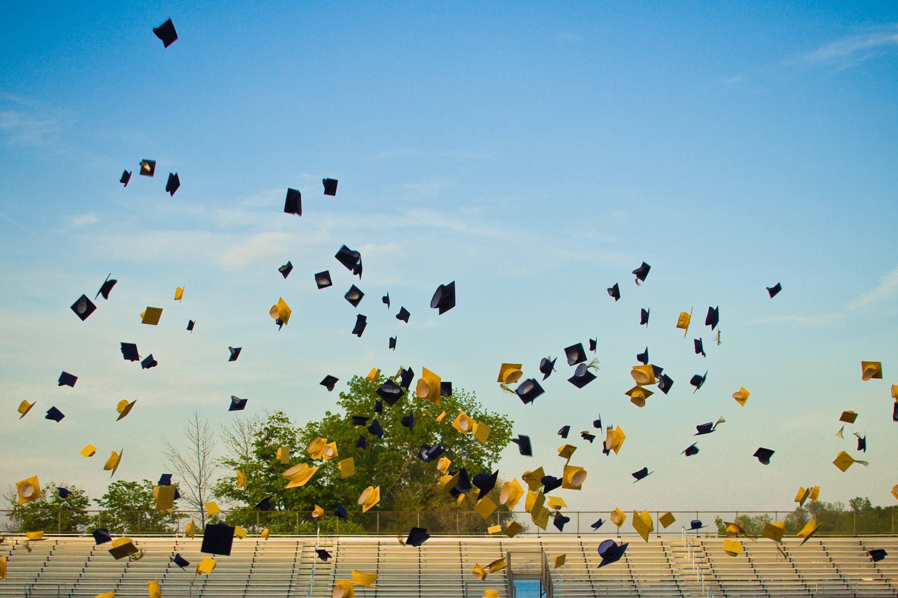 Blue and yellow graduation caps are in the air with a stadium and and lights in the background.