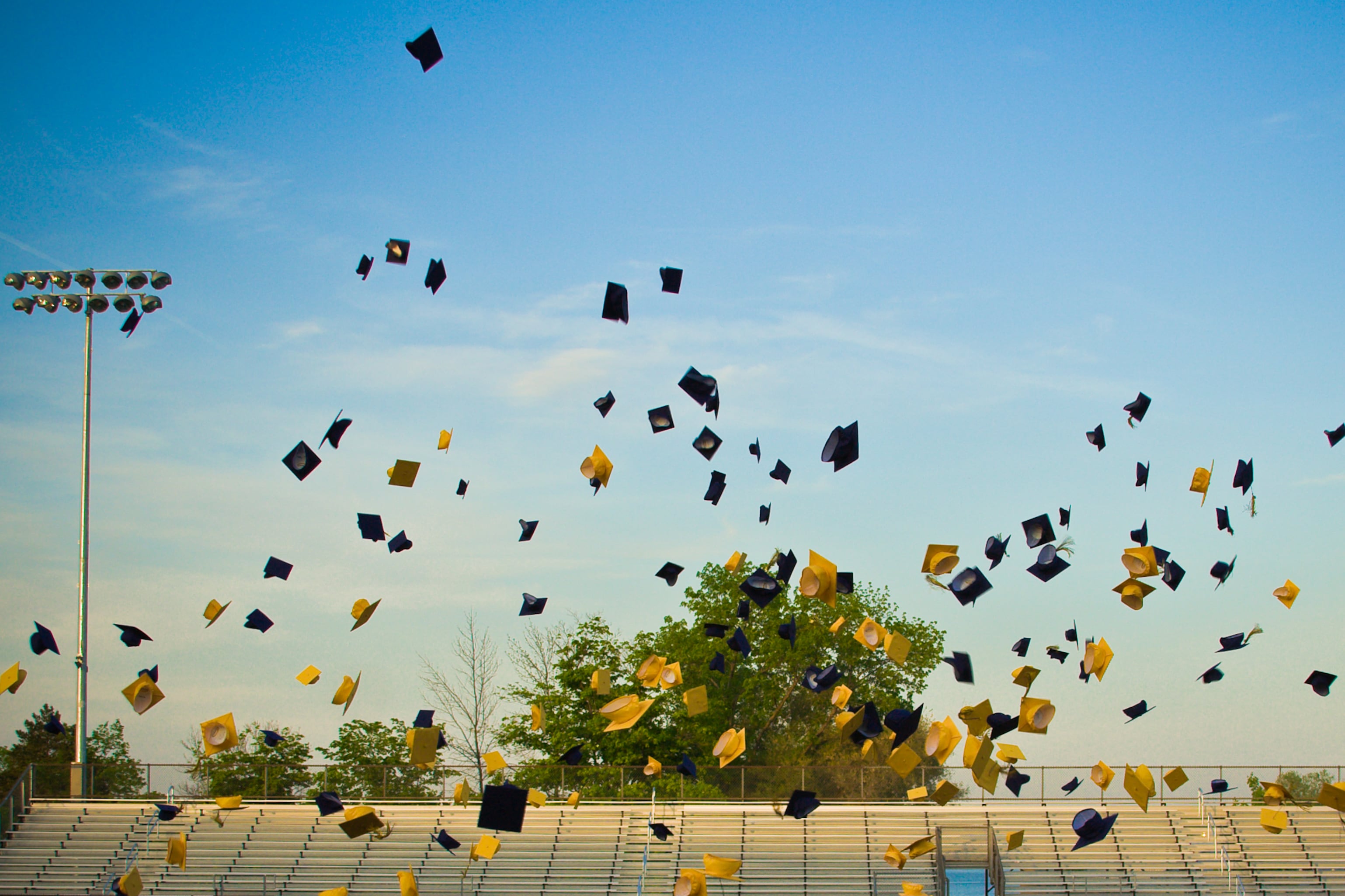 Blue and yellow graduation caps are in the air with a stadium and and lights in the background.