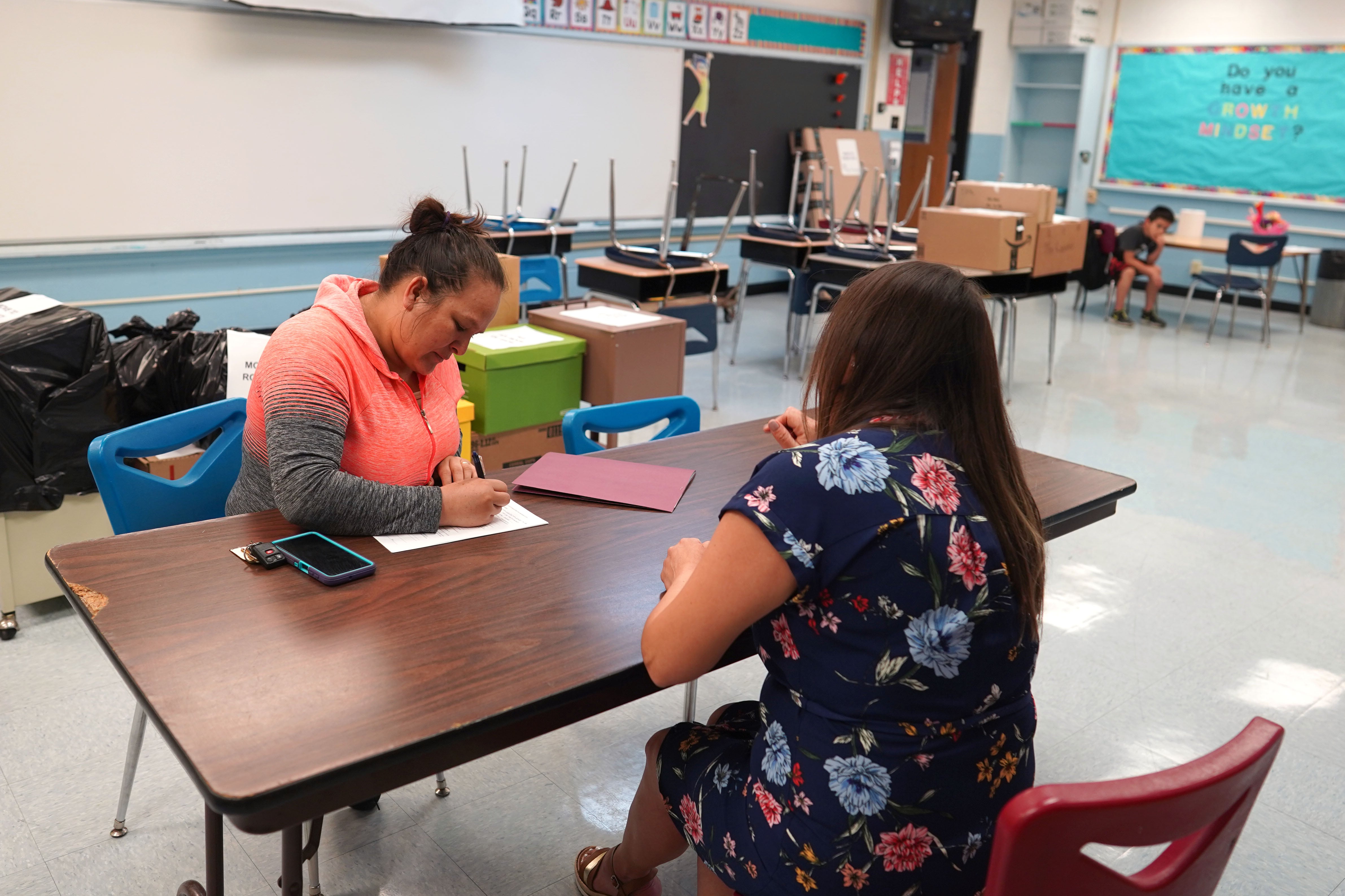 Two adult women sit at a long table inside a school classroom.