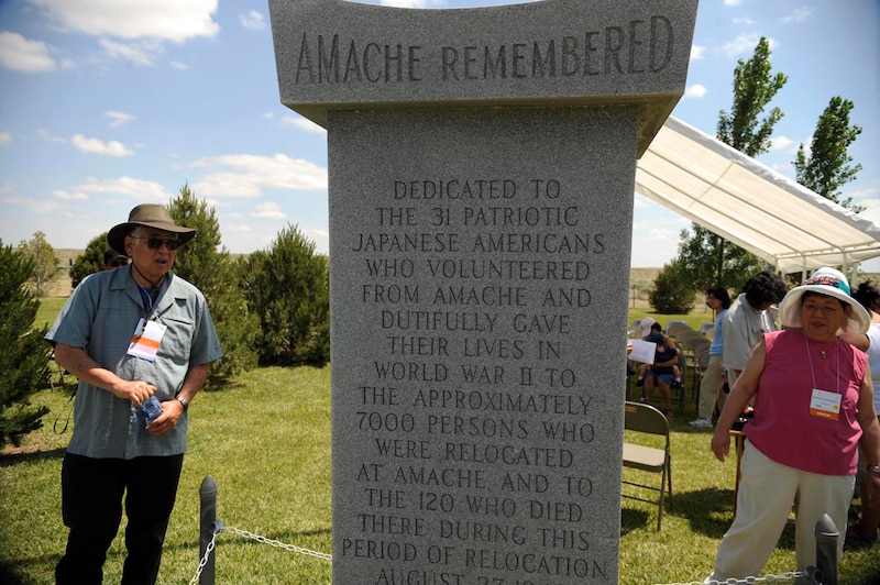 A photograph of two adults standing on each side of a large stone memorial statue outside on a sunny day.
