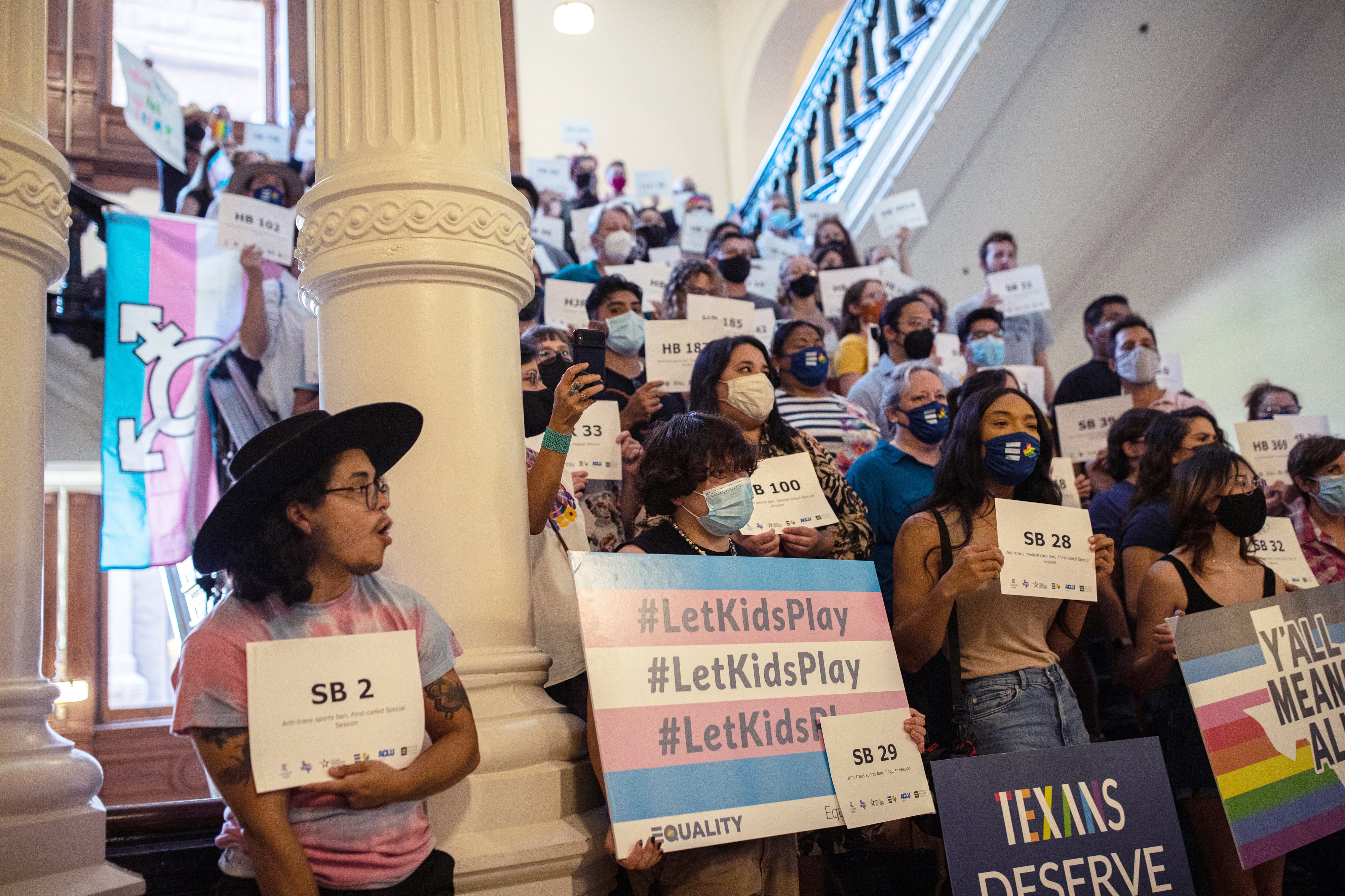 A group of people with protest signs stand on steps inside a white building with a large white column in the front left