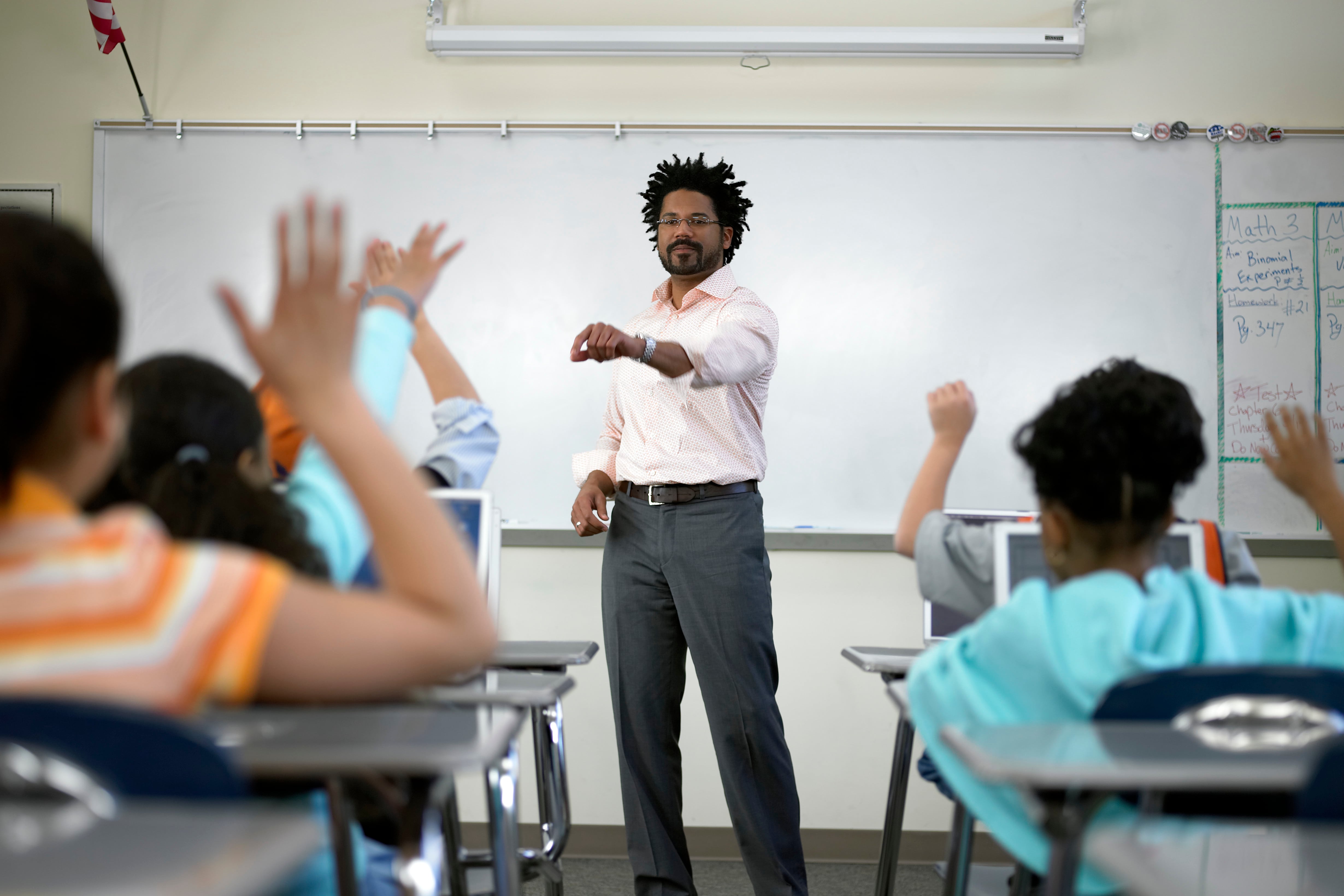 A male teacher wearing an off-white shirt and dark pants stands in front of a white board and a row of students sitting at their desks.