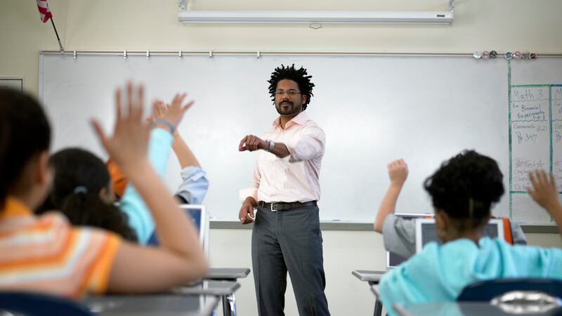 A male teacher wearing an off-white shirt and dark pants stands in front of a white board and a row of students sitting at their desks.
