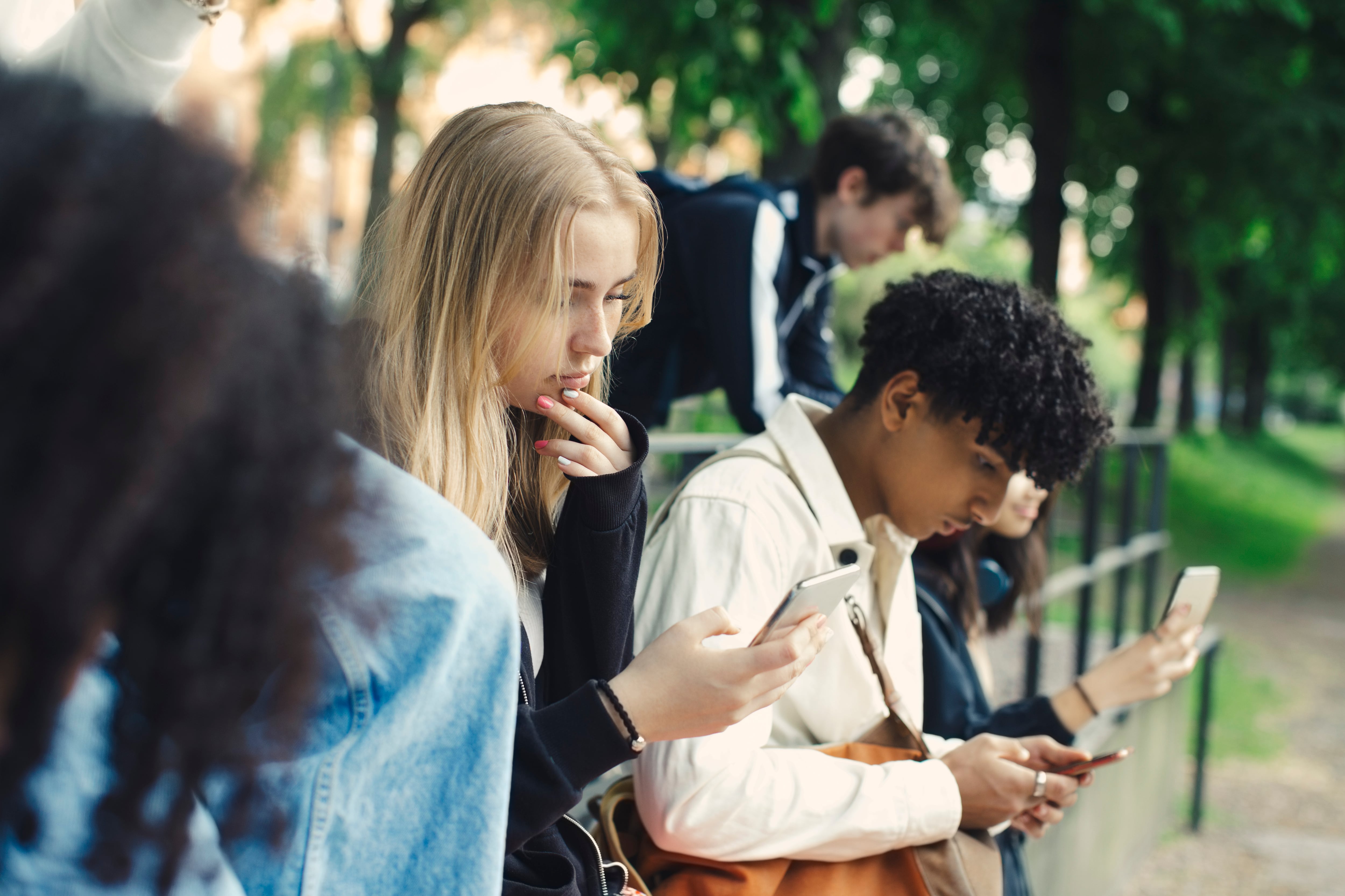 Three students stand outdoors looking down at their phones.