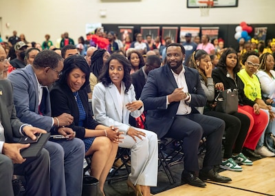 Rows of people sit in chairs in a school gym.