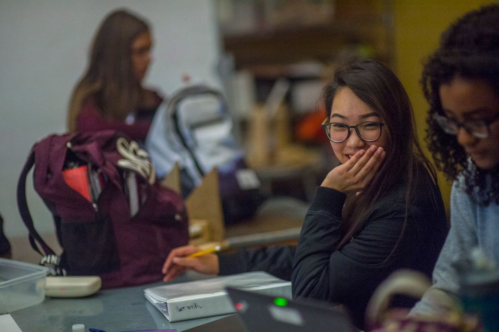 Two high school students work at a desk with one student in the background.