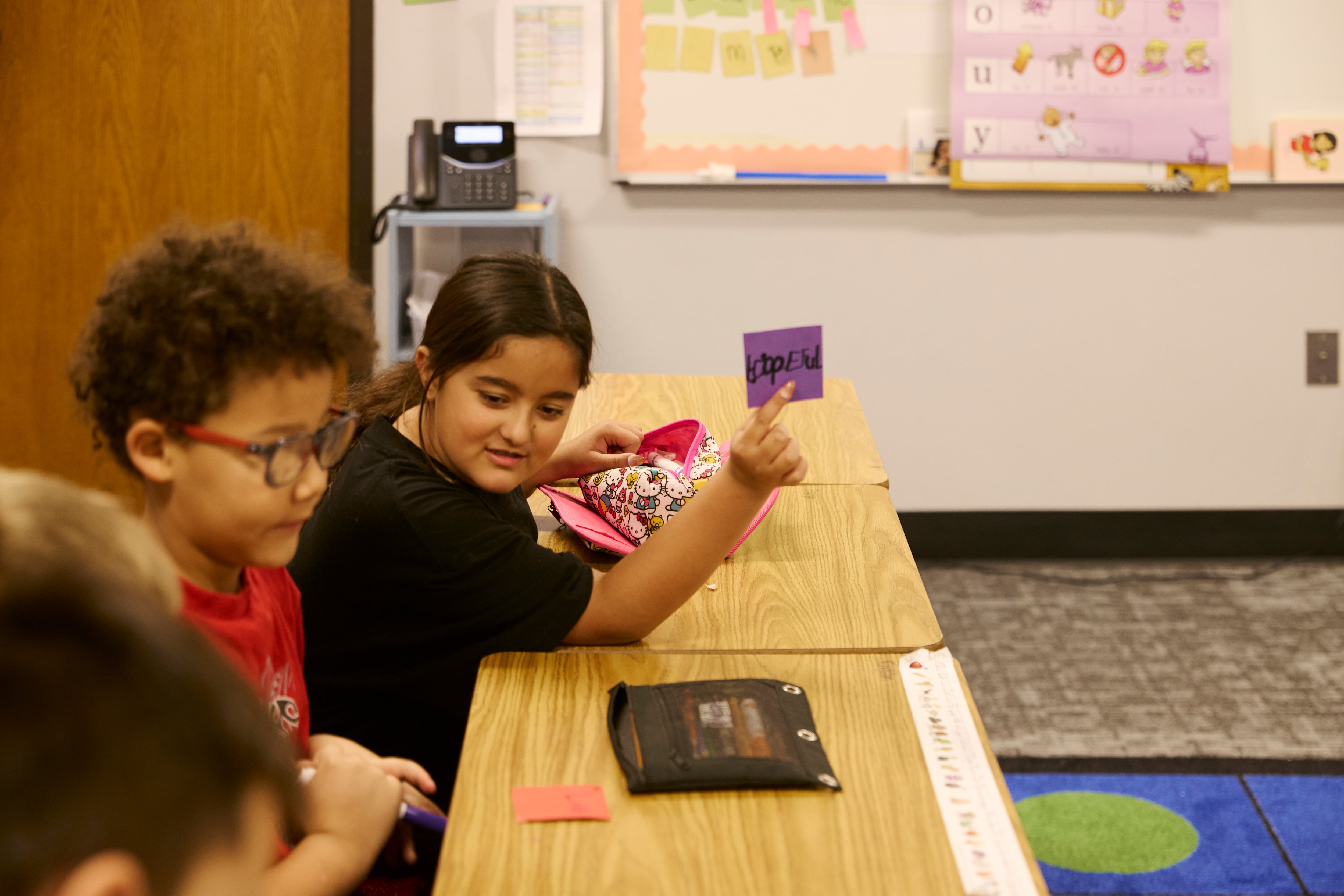 Two elementary students sit at a wooden desk in a colorful classroom.