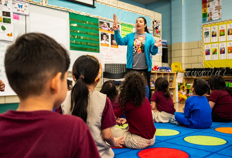An adult teacher wearing a blue sweater stands in front of a group of young students sitting on a colorful rug in an elementary class.