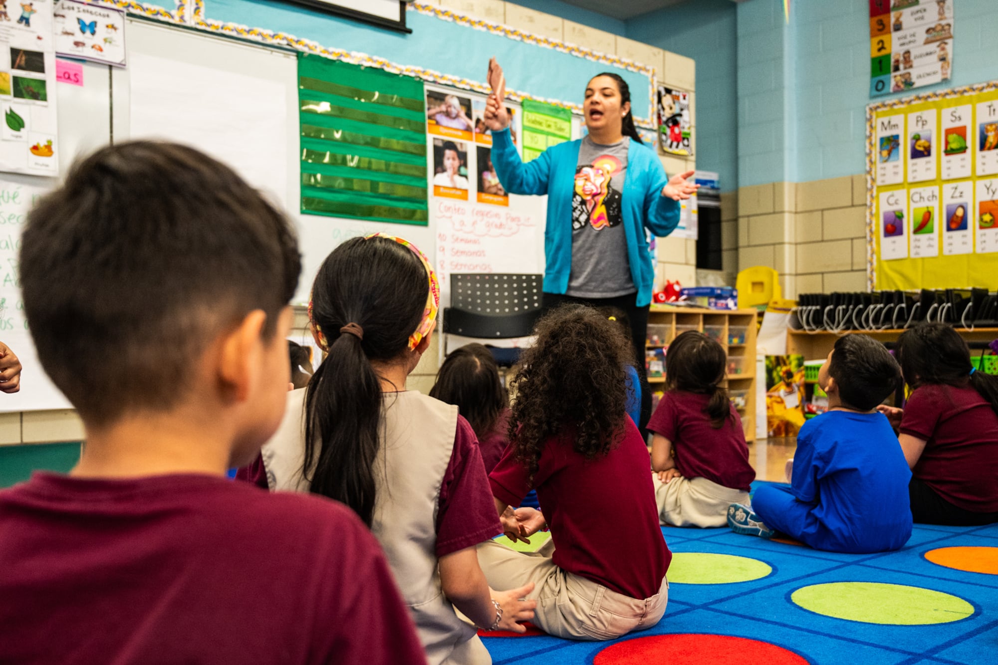 An adult teacher wearing a blue sweater stands in front of a group of young students sitting on a colorful rug in an elementary class.