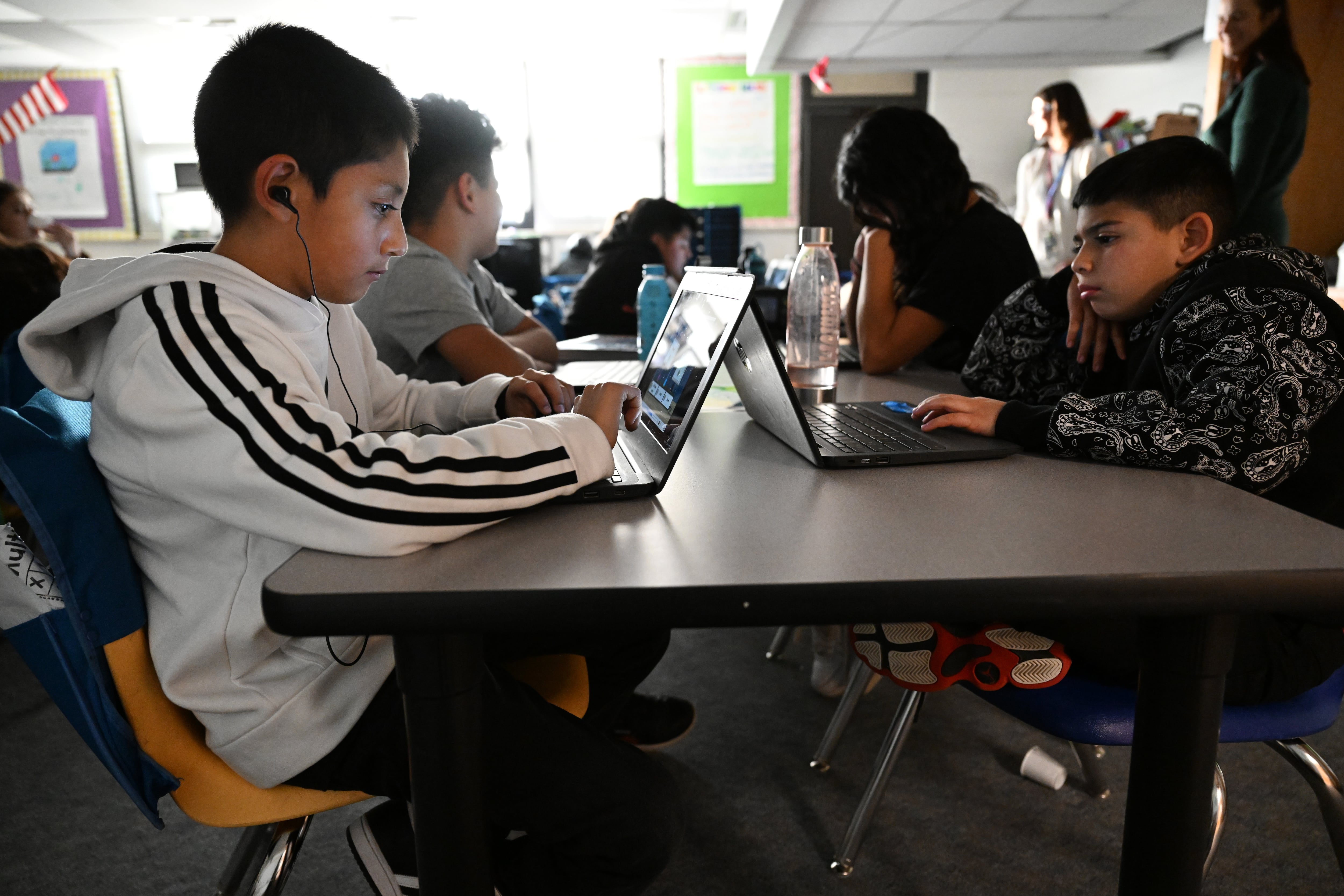 Students sit working on laptops on a large desk. Light from a class window comes in behind them.