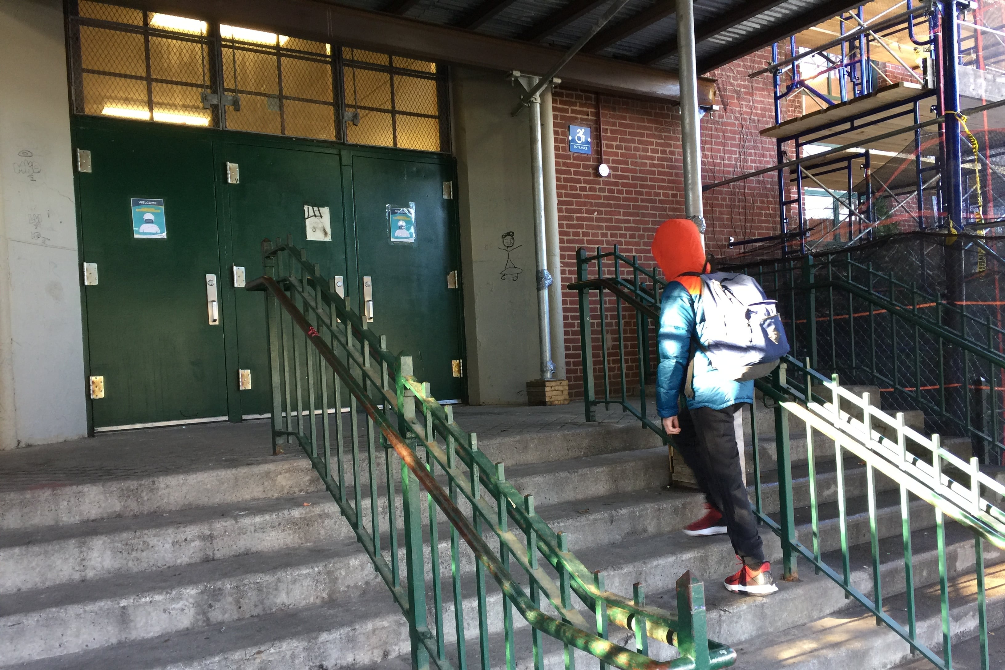 A student in a backpack walks up concrete steps toward green double doors at a school.