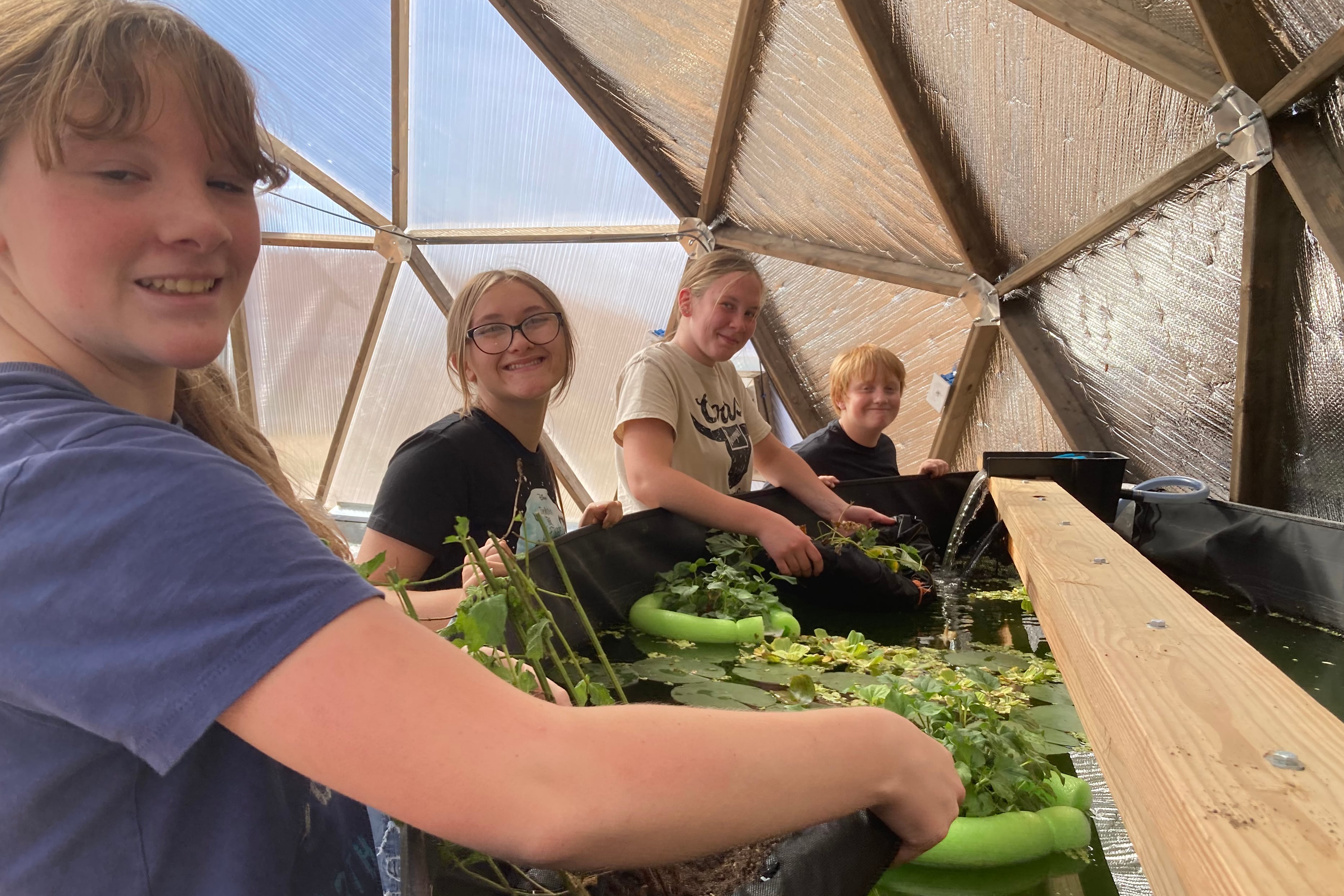 A photograph of four students posing for a photograph next to a large tub of water with greenery.