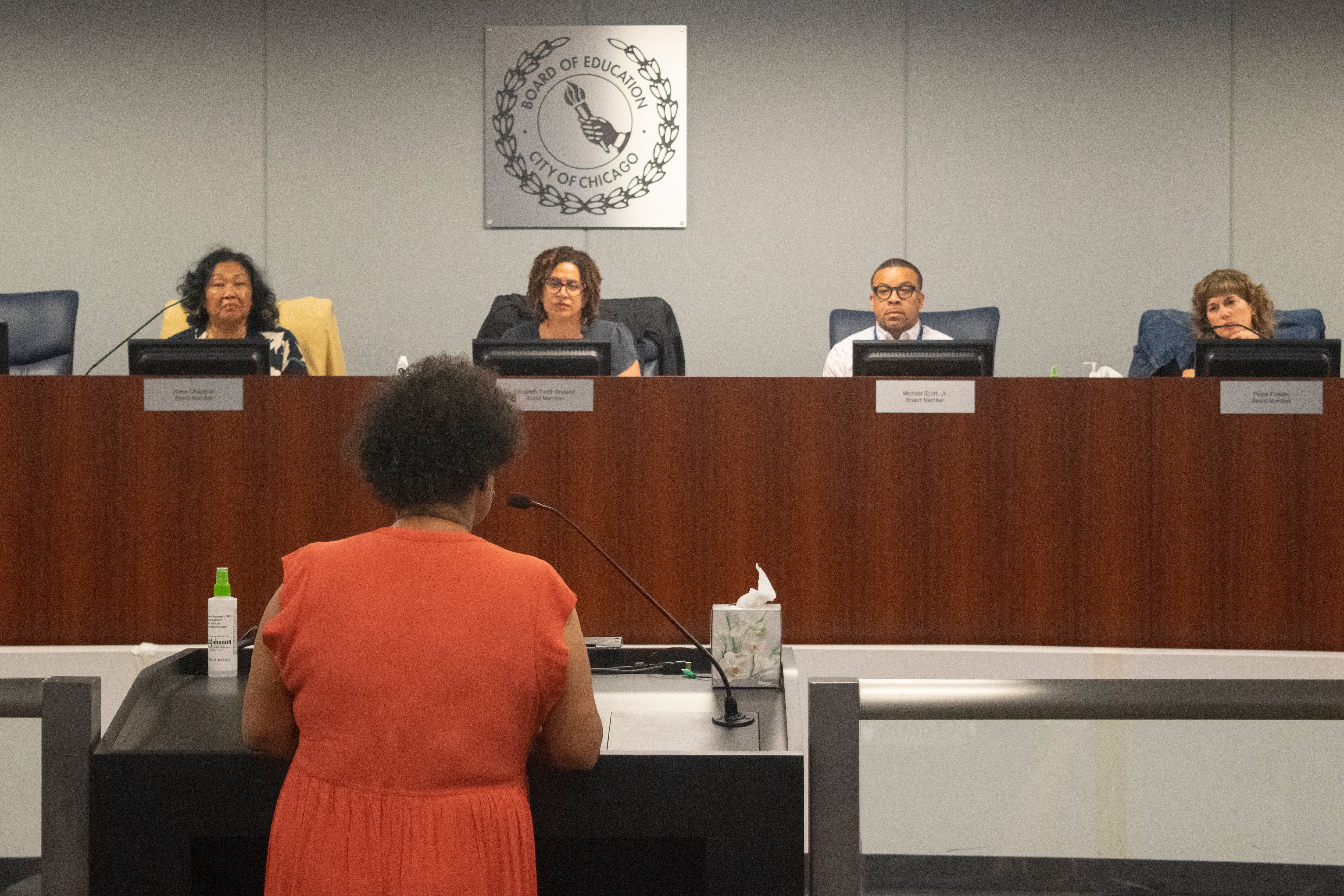 A woman speaks at a lectern at a Chicago school board meeting. Her back is to the camera, with her face turned toward the members of the school board, who sit at a shared dais.