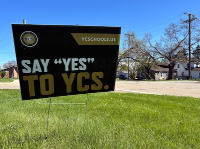 A black yard sign with yellow and white letters on a grassy lawn with a house in the background.