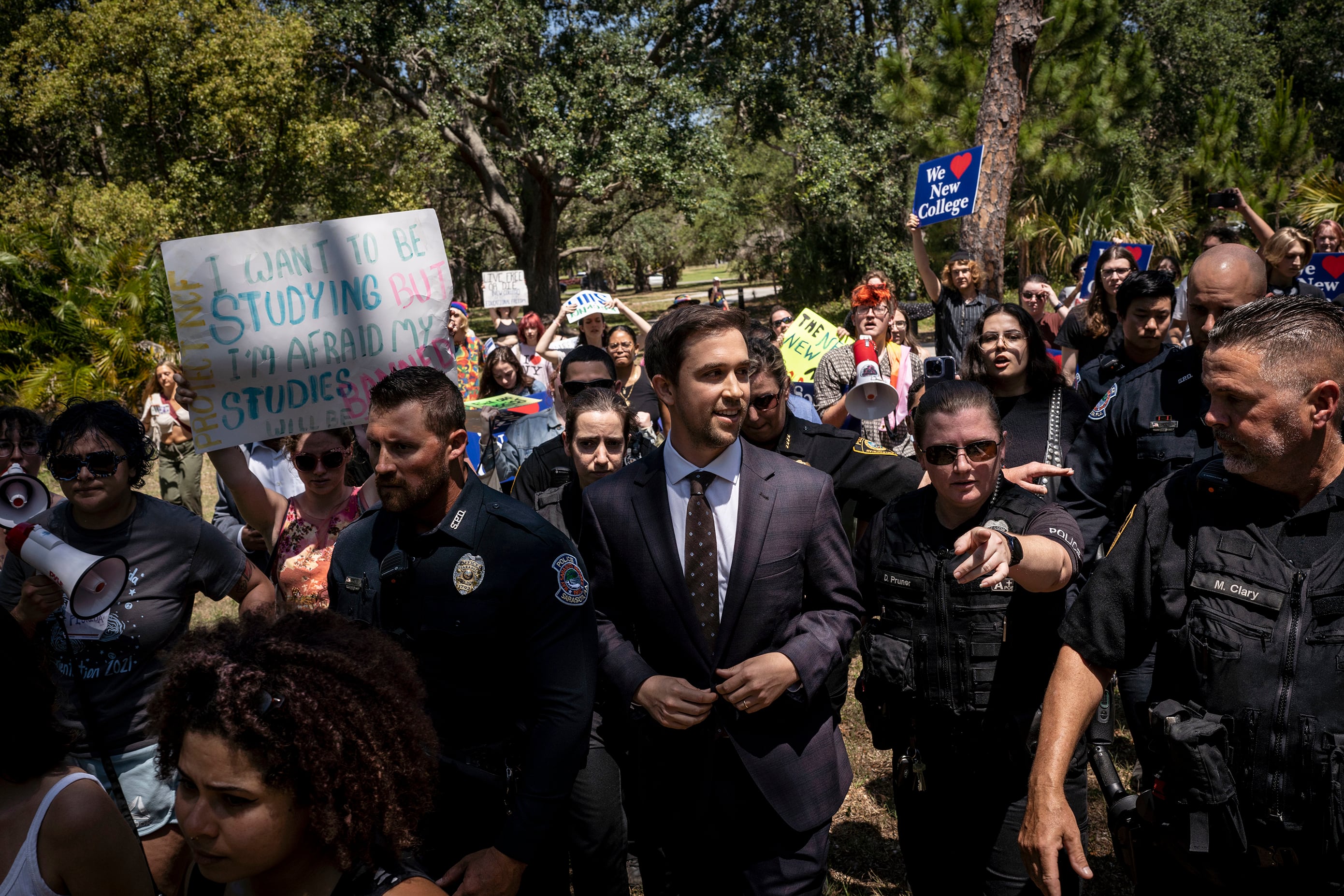 A crowd of people including a man in a suit, police officers and protestors in front of a wall of green trees.