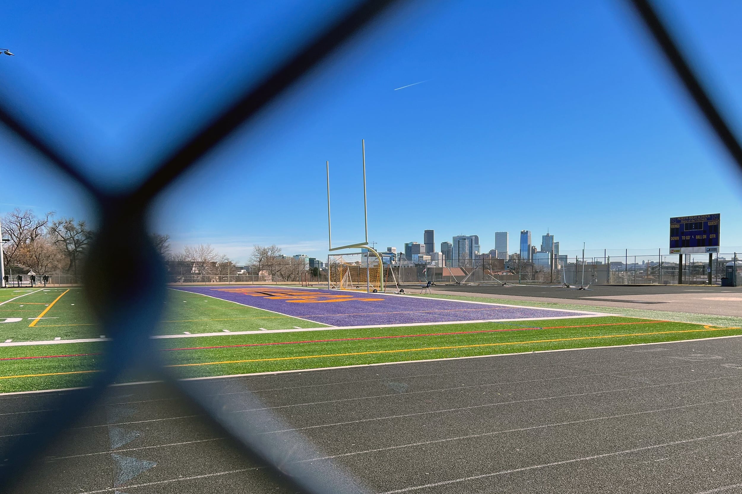 A skyline of Denver, Co is seen through a wire fence in the foregound and a track and field in the middleground.
