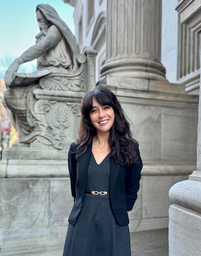 A white woman with medium length brown hair stands posing for a portrait near a stone column and statue.