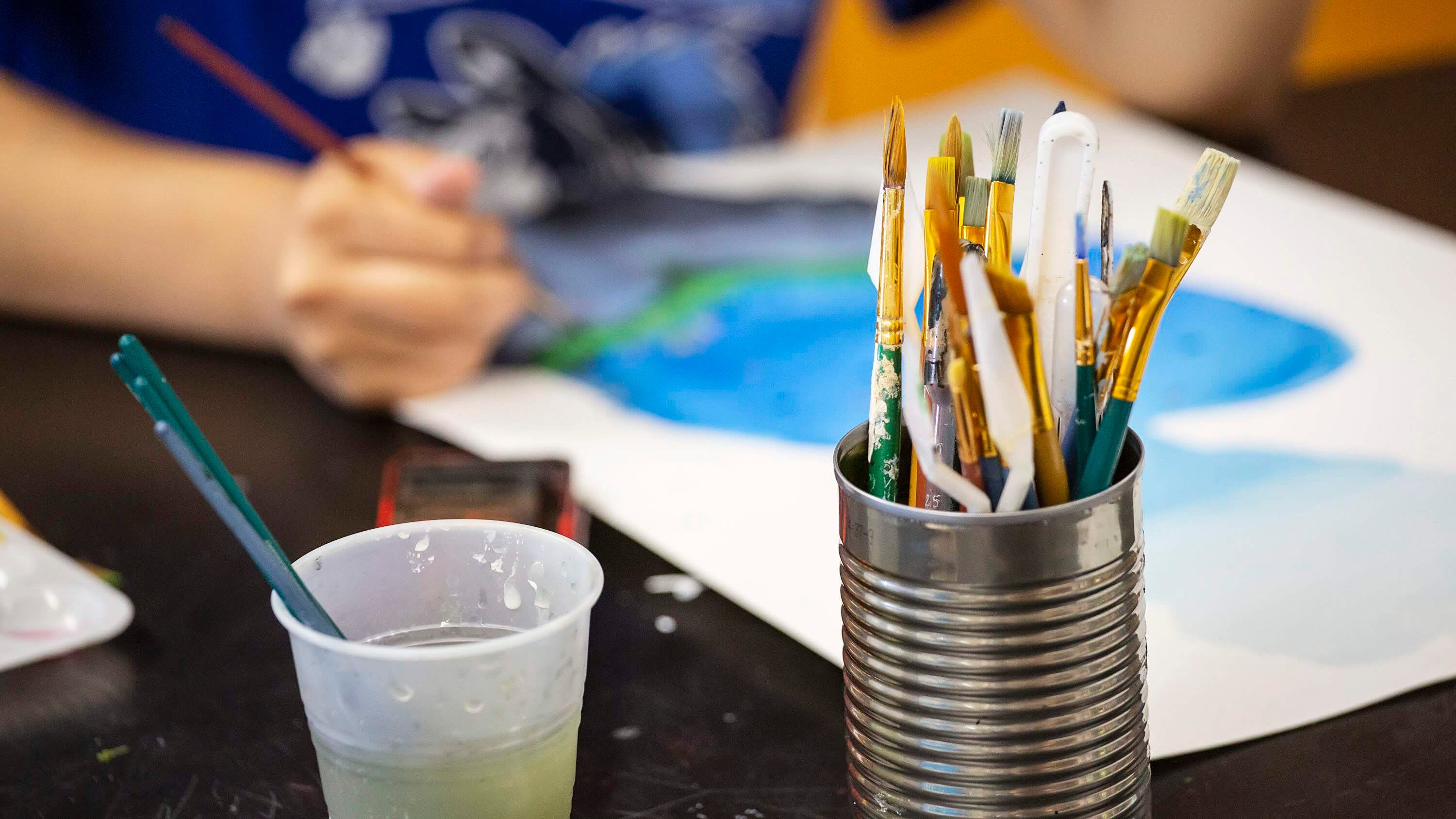 A close-up photo of a tin can full of paintbrushes. In the background, a student paints in art class.