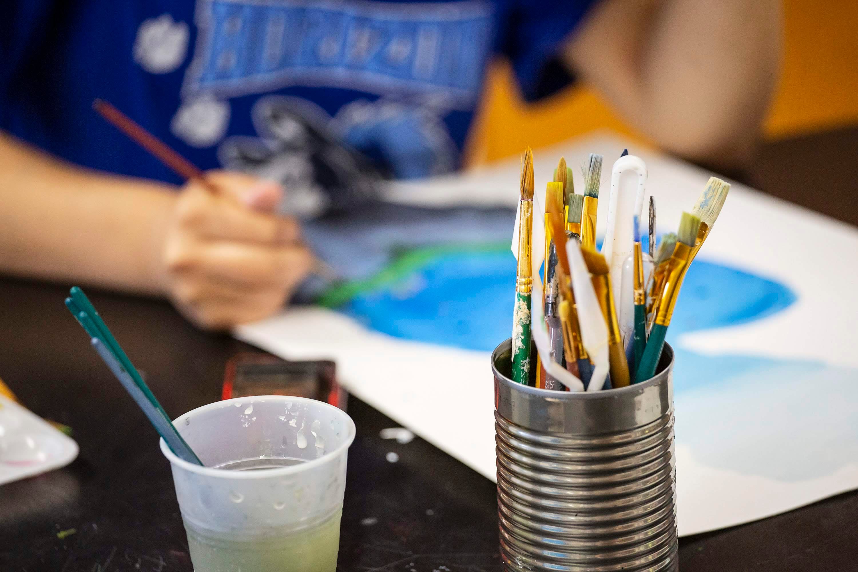 A close-up photo of a tin can full of paintbrushes. In the background, a student paints in art class.