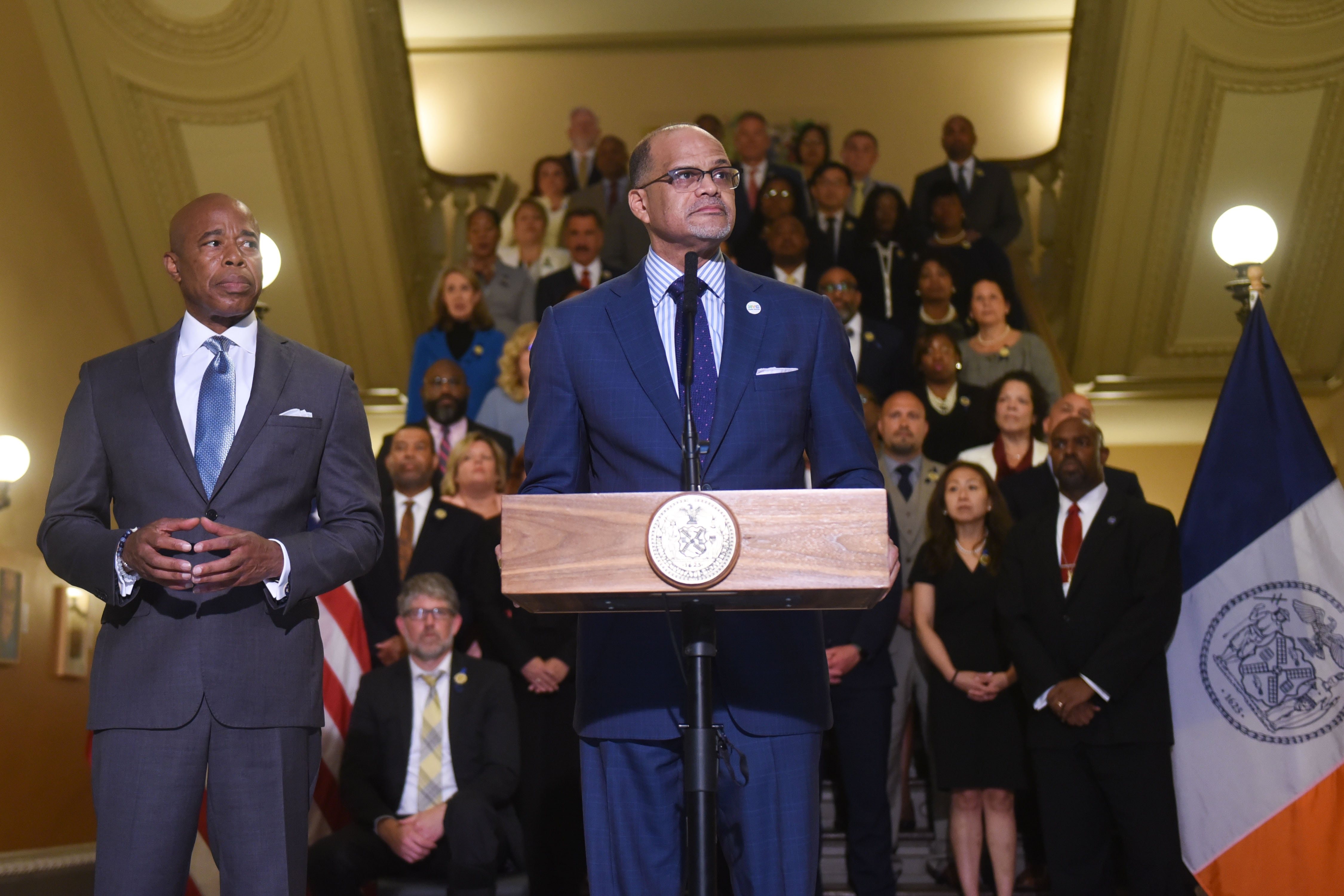 A man in a blue suit stands at a lectern flanked by another man, with dozens of people on stairs behind them.