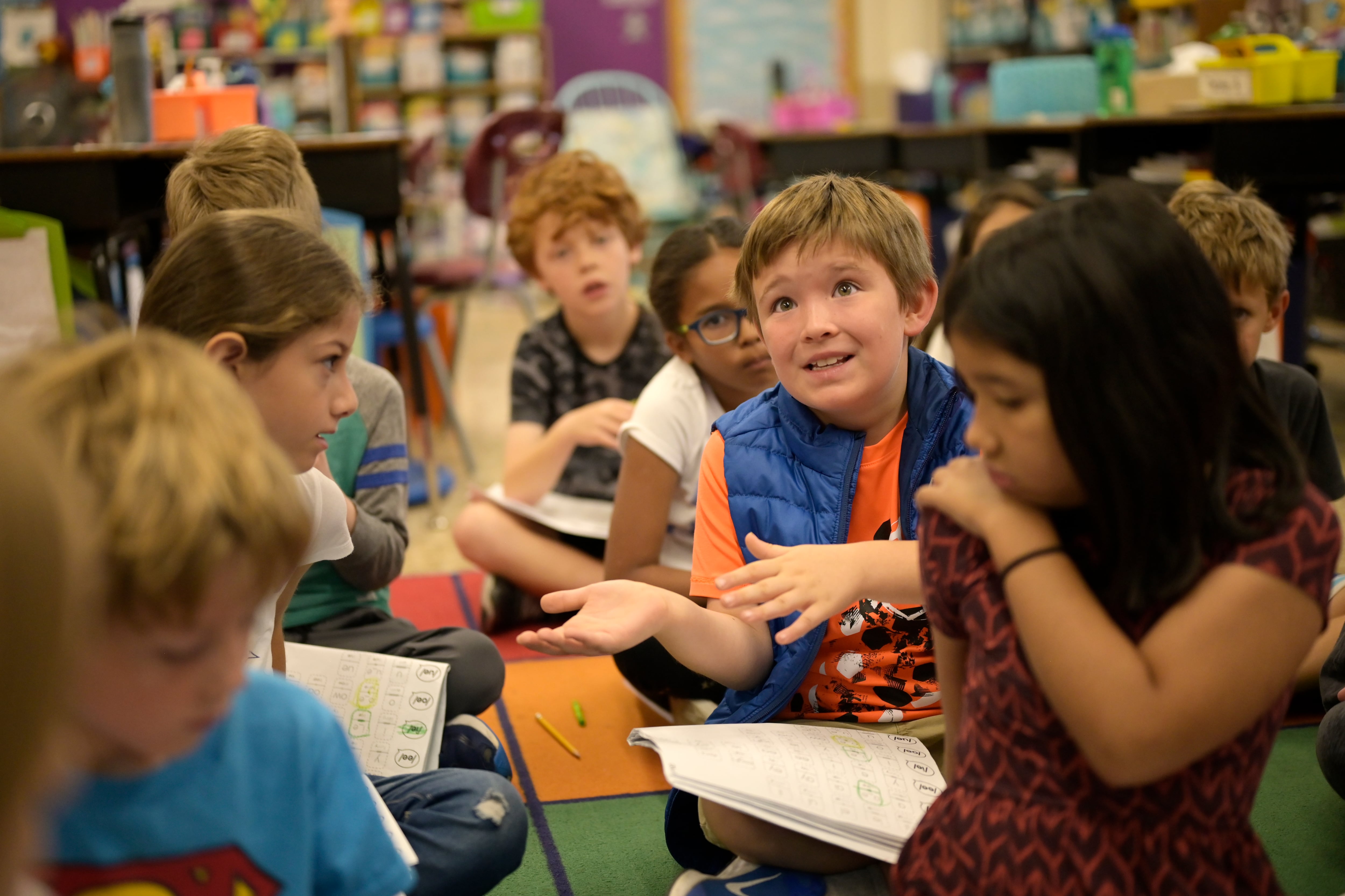 A boy with an orange shirt and blue vests sits on the classroom carpet with other second-grade students during a reading lesson.