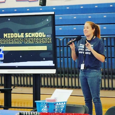 A woman with a ponytail and wearing a blue shirt and blue jeans speaks at a middle school leadership conference.
